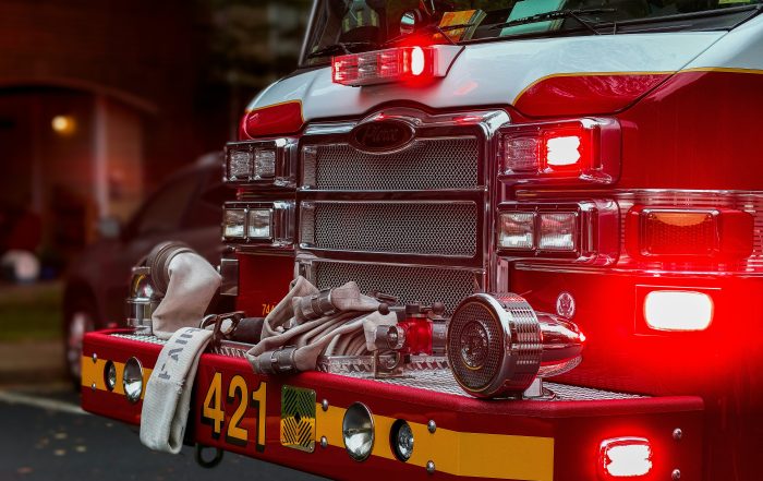 A photo of the front of a fire truck with it's lights on and a white fire hose laying across the front bumper.