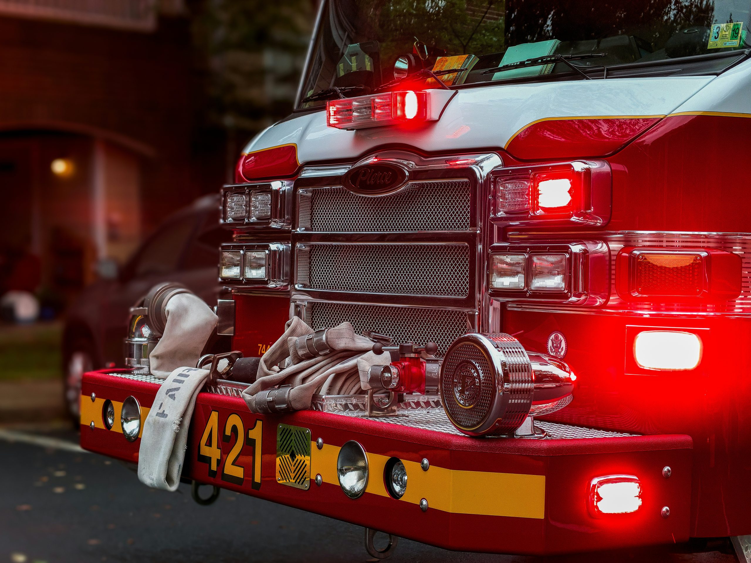 obi-HMcd1bWLTjo-unsplash A photo of the front of a fire truck with it's lights on and a white fire hose laying across the front bumper.