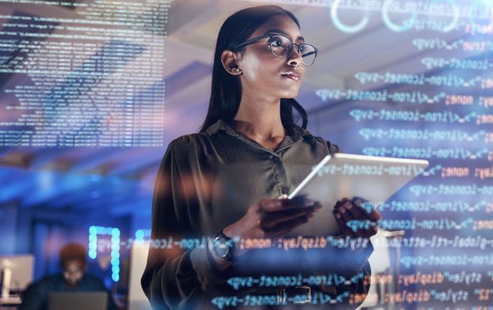 A woman wearing glasses holds a tablet in her hand and looks at computer data sets in front of her.